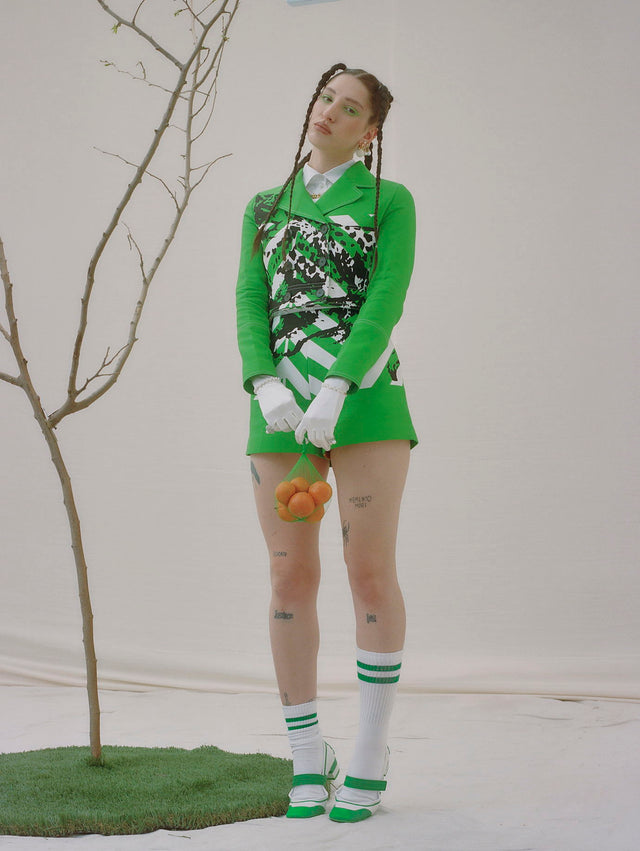 Woman in a green outfit holding an orange beside a bare tree