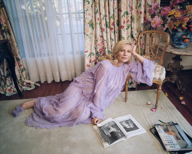 Woman in a lavender dress reclining on a rug beside an open magazine in a floral room